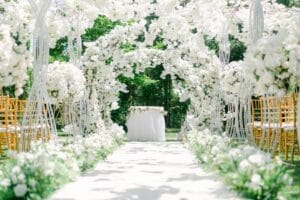An elegant outdoor wedding venue setup with white floral arches and chairs.