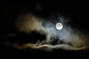 A full moon illuminates the night sky, shrouded by eerie clouds creating a spooky atmosphere.